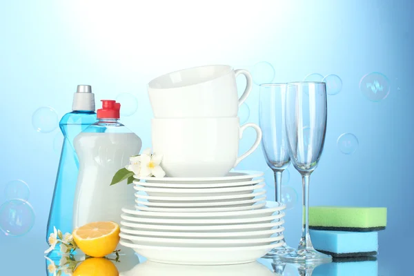 Piatti puliti vuoti, bicchieri e tazze con liquido di lavaggio piatti, spugne e limone su sfondo blu — Foto Stock