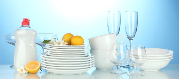 Empty clean plates, glasses and cups with dishwashing liquidand lemon on blue background