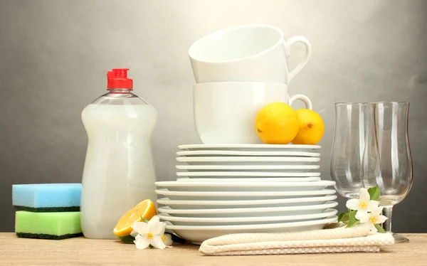 Άδειο καθαρά πιάτα, ποτήρια και κύπελλα με υγρό πιάτων, σφουγγάρια και λεμόνι σε ξύλινο τραπέζι σε γκρι φόντο — Φωτογραφία Αρχείου