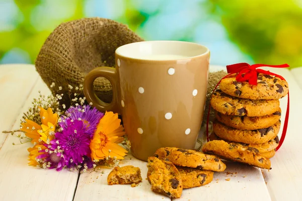 Cup of milk, chocolate chips cookies with red ribbon and wildflowers on wooden table on green background — Stock Photo, Image