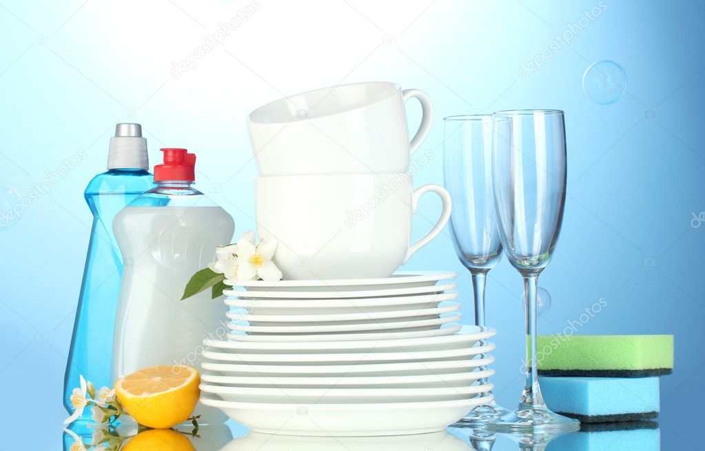 Empty clean plates, glasses and cups with dishwashing liquid, sponges and lemon on blue background