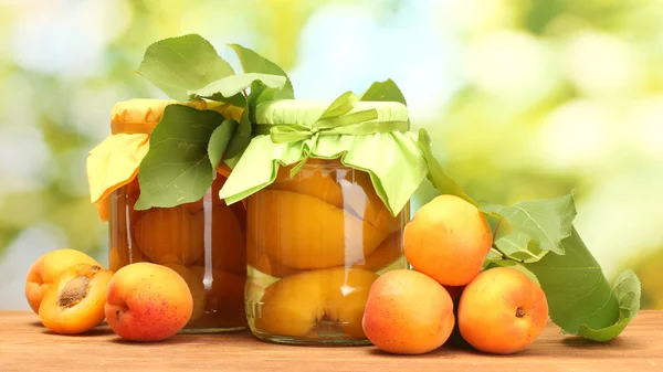 Canned apricots in a jars and sweet apricots on wooden table on green background — Stock Photo, Image