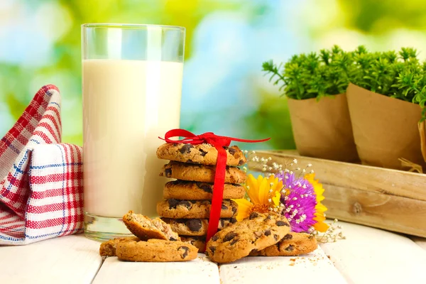 Sklenici mléka, čokoládová čipy cookies s červenou stužku a květy na dřevěný stůl na zeleném pozadí — Stock fotografie