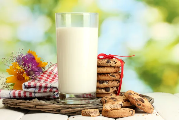 Szklanki mleka, czekoladowe żetony ciasteczka z czerwoną wstążką i kwiaty na drewnianym stole na zielonym tle — Zdjęcie stockowe