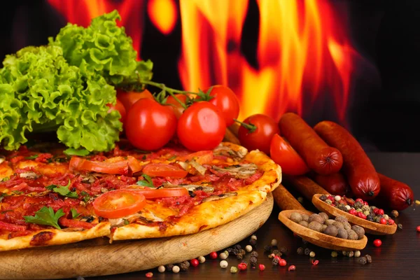 Deliciosa pizza, salami, tomates y especias en mesa de madera sobre fondo de llama —  Fotos de Stock