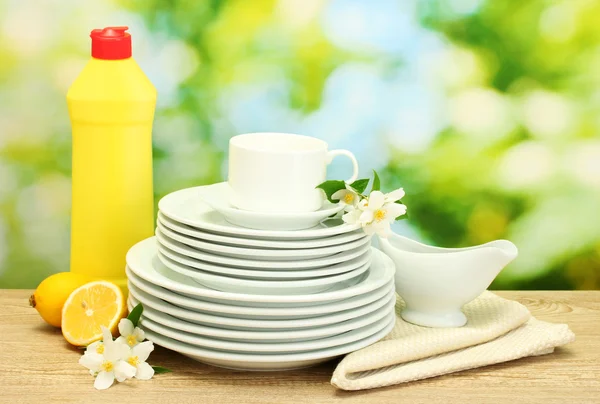 Assiettes et tasses propres vides avec liquide vaisselle, fleurs et citron sur table en bois sur fond vert — Photo