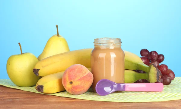 Frasco com alimentos para bebês de frutas, colher e frutas em guardanapo colorido no fundo azul — Fotografia de Stock