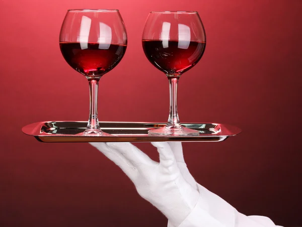 Mano in guanto contenente vassoio d'argento con occhiali da vino su sfondo rosso — Foto Stock