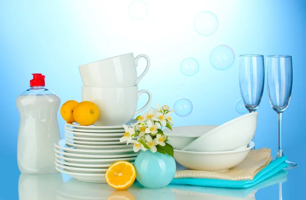 Platos limpios vacíos, vasos y tazas con líquido lavavajillas y limón sobre fondo azul —  Fotos de Stock