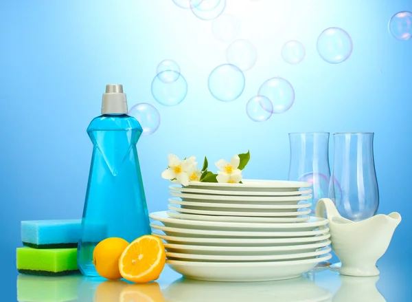 Piatti puliti vuoti e bicchieri con detersivo per piatti, spugne e limone su fondo blu — Foto Stock