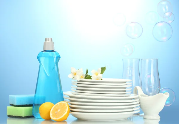 Άδειο καθαρά πιάτα και ποτήρια με υγρό πιάτων, σφουγγάρια και λεμόνι σε μπλε φόντο — Φωτογραφία Αρχείου
