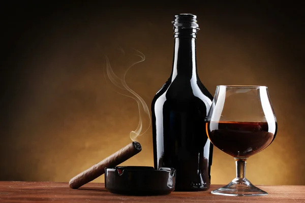 Botella y vaso de brandy y cigarro sobre mesa de madera sobre fondo marrón — Foto de Stock