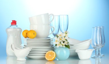 Empty clean plates, glasses and cups with dishwashing liquid and lemon on blue background clipart