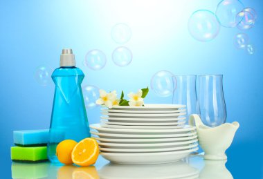 Empty clean plates and glasses with dishwashing liquid, sponges and lemon on blue background clipart