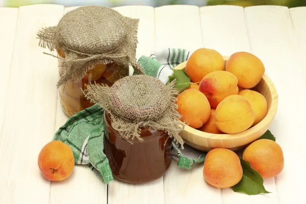 Консервированные абрикосы и варенье в банках и спелые абрикосы в миске на белом деревянном столе — стоковое фото