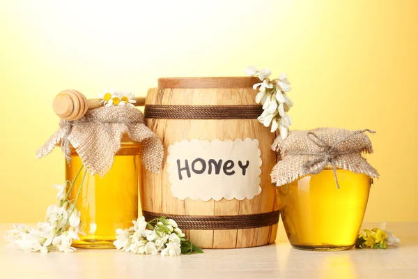 Miel dulce en barril y frascos con flores de acacia sobre mesa de madera sobre fondo amarillo —  Fotos de Stock