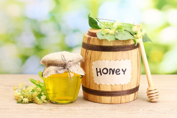 Jar and barrel with linden honey and flowers on wooden table on green background — Stock Photo, Image