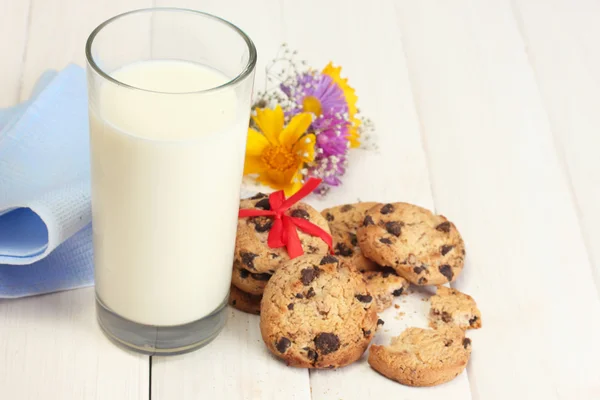 Sklenici mléka, čokoládová čipy cookies s červenou stužku a květy na dřevěný stůl — Stock fotografie