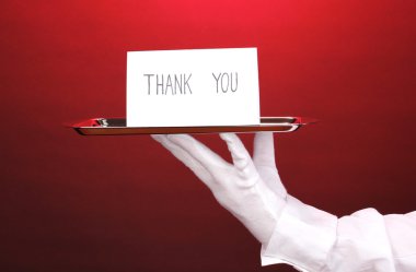 Hand in glove holding silver tray with card saying thank you on red background clipart