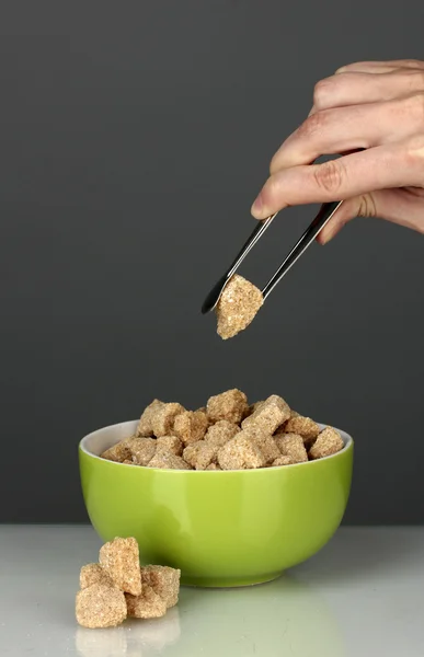 Hnědý třtinový cukr kostky v misce a Zenske ruce drží kleštičky na cukr na šedém pozadí — Stock fotografie