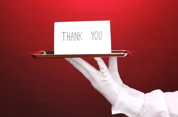Guante de mano sosteniendo bandeja de plata con tarjeta diciendo gracias sobre fondo rojo — Foto de Stock