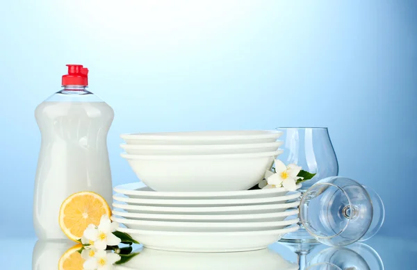 Platos y vasos limpios vacíos con líquido lavavajillas y limón sobre fondo azul —  Fotos de Stock