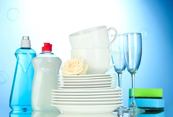 Platos limpios vacíos, vasos y tazas con líquido lavavajillas y esponjas sobre fondo azul —  Fotos de Stock