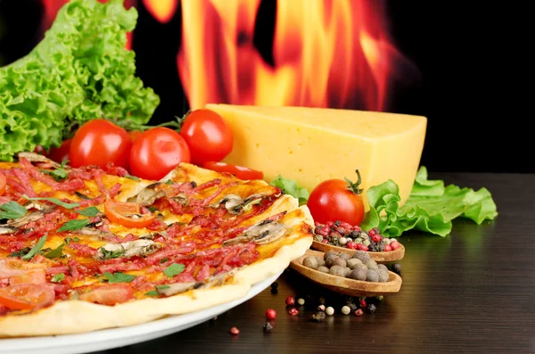 Herkullinen pizza, salami, tomaatit ja mausteet puupöydällä liekkitaustalla — kuvapankkivalokuva