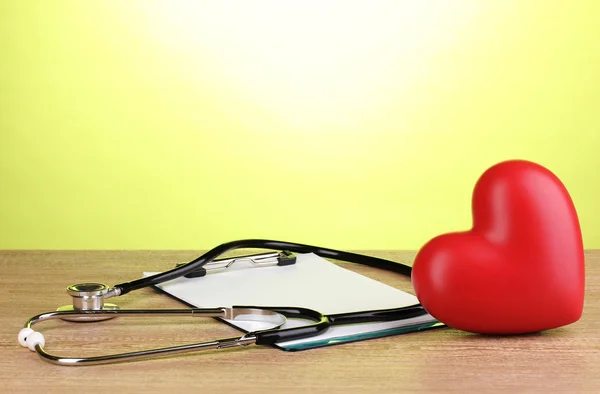 クリップボードと緑の背景に木製のテーブルの上の心と医療の聴診器 — ストック写真