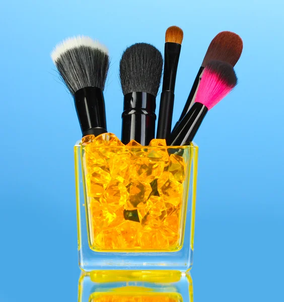Pincéis de maquiagem em copo de vidro com pedras no fundo azul — Fotografia de Stock