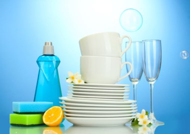 Empty clean plates, glasses and cups with dishwashing liquid, sponges and lemon on blue background clipart