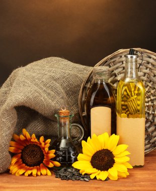 Oil in bottles, sunflowers and seeds, on wooden table on brown background clipart