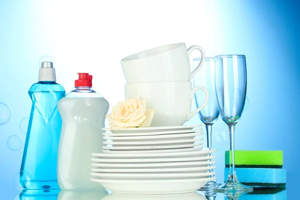 Platos limpios vacíos, vasos y tazas con líquido lavavajillas y esponjas sobre fondo azul —  Fotos de Stock