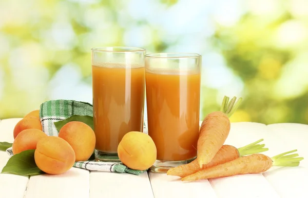 Gläser mit Karotten und Aprikosensaft auf weißem Holztisch auf grünem Hintergrund — Stockfoto