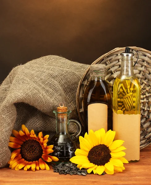 Oil in bottles, sunflowers and seeds, on wooden table on brown background — Stock Photo, Image