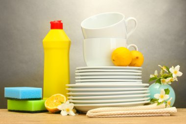 Empty clean plates, glasses and cups with dishwashing liquid, sponges and lemon on wooden table on grey background clipart