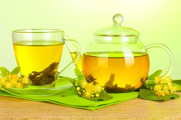 Teapot and cup with linden tea and flowers on wooden table on green background — Stock Photo, Image