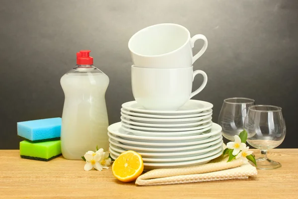 Pratos limpos vazios, copos e copos com líquido de lavar louça, esponjas e limão na mesa de madeira em fundo cinza — Fotografia de Stock