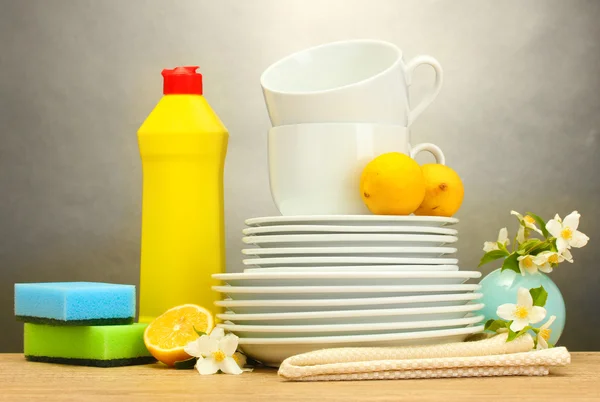 Пустые чистые тарелки, стаканы и чашки с жидкостью для мытья посуды, губки и лимон на деревянном столе на сером фоне — стоковое фото
