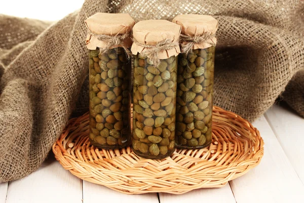 Glass jars with tinned capers in sack on white wooden background — Stock Photo, Image
