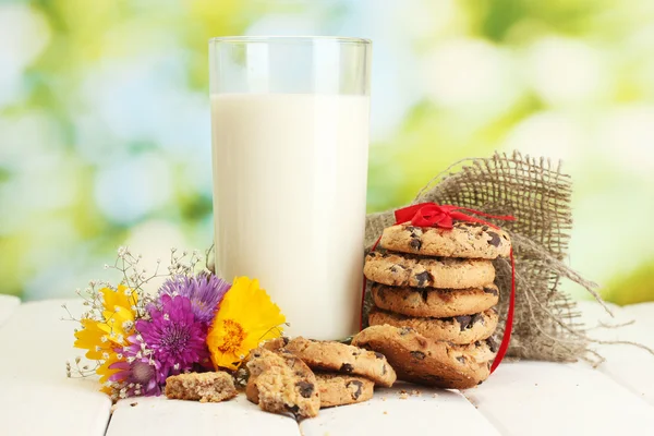 Glas mjölk, choklad chip cookies med rött band och blommor på träbord på grön bakgrund — Stockfoto