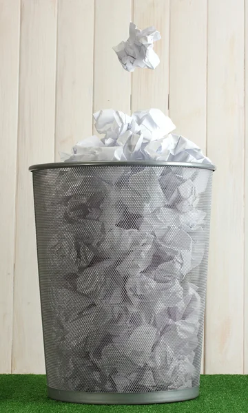 Metal trash bin from paper on grass on wooden background