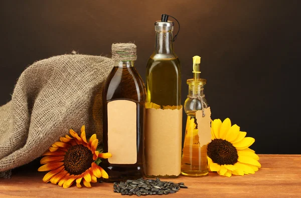 Oil in bottles, sunflowers and seeds, on wooden table on brown background — Stock Photo, Image