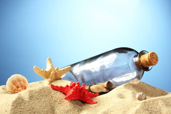 Skleněná láhev s poznámkou uvnitř na písku, na modrém pozadí — Stock fotografie