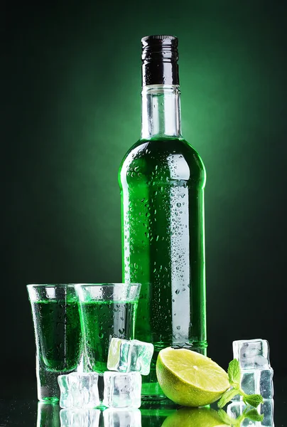 Bottiglia e bicchieri di assenzio con calce e ghiaccio su fondo verde — Foto Stock