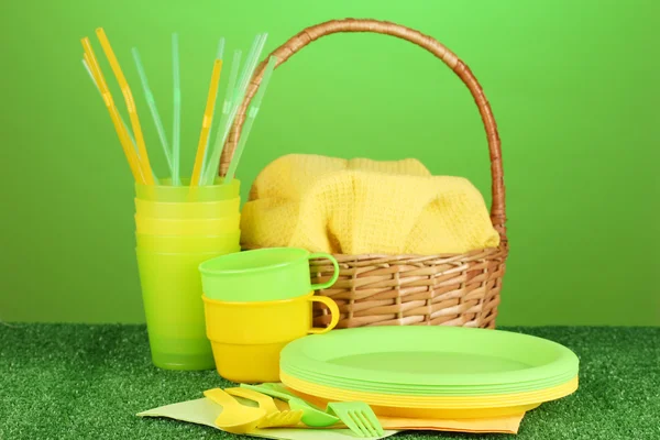 Plástico brilhante utensílios de mesa descartáveis e cesta de piquenique no gramado em fundo colorido — Fotografia de Stock