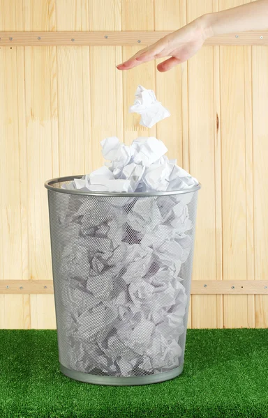 Hand going garbage in metal trash bin from paper on grass on wooden background — Stock Photo, Image