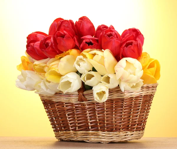 Krásné tulipány v koši na dřevěný stůl na žlutém podkladu — Stock fotografie