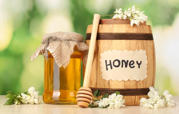 Γλυκό μέλι στο βαρέλι και βάζο με λουλούδια της ακακίας στο ξύλινο τραπέζι σε πράσινο φόντο — Φωτογραφία Αρχείου