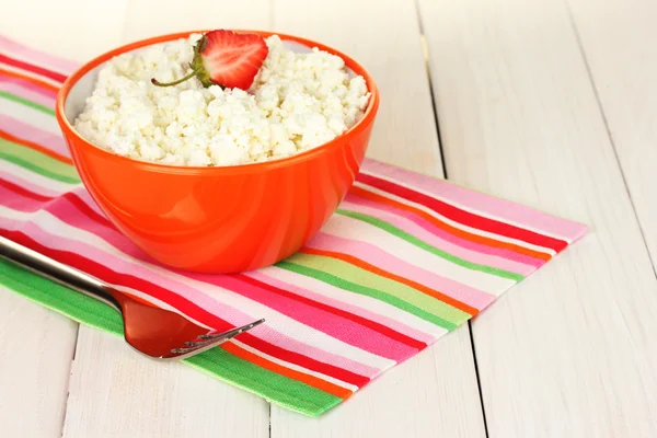 Cottage cheese with strawberry in orange bowl and fork on colorful napkin on white wooden table close-up
